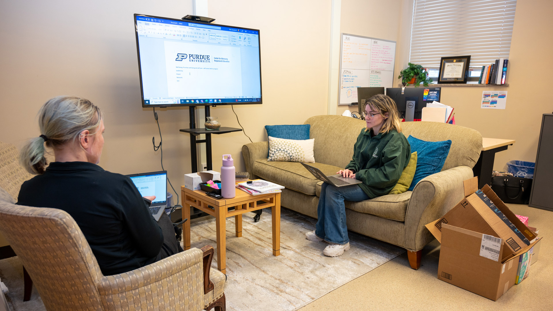 Two individuals are working in a cozy office setting; one is using a laptop seated on an armchair while the other is working on a laptop placed on a standing desk, with a Purdue University document open on the screen.